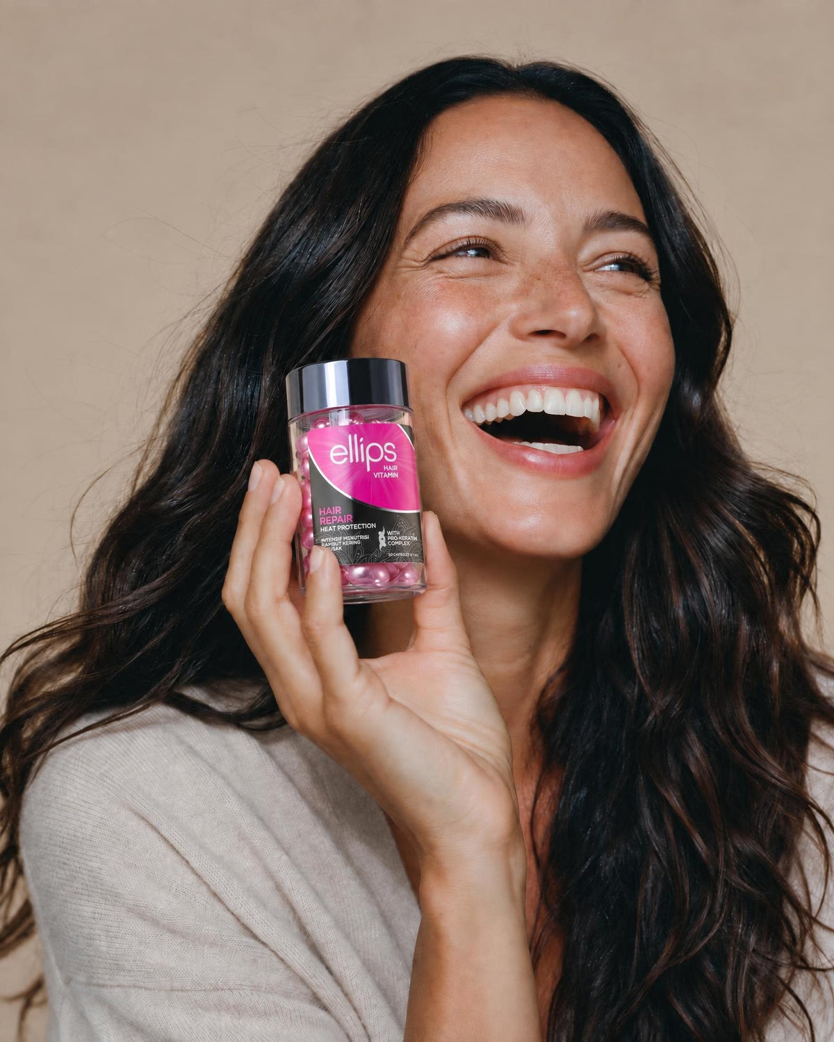 Naurava nainen, jolla on pitkät tummat hiukset, pitelee Ellips Hair Repair -purkkia