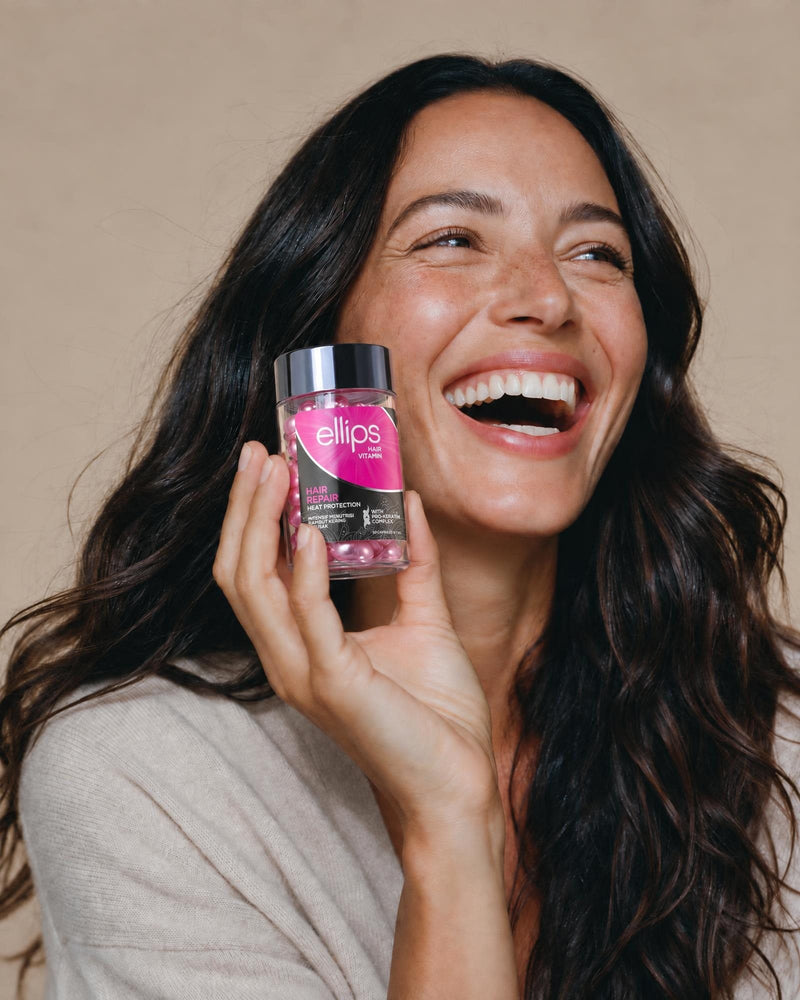 Naurava nainen, jolla on pitkät tummat hiukset, pitelee Ellips Hair Repair -purkkia