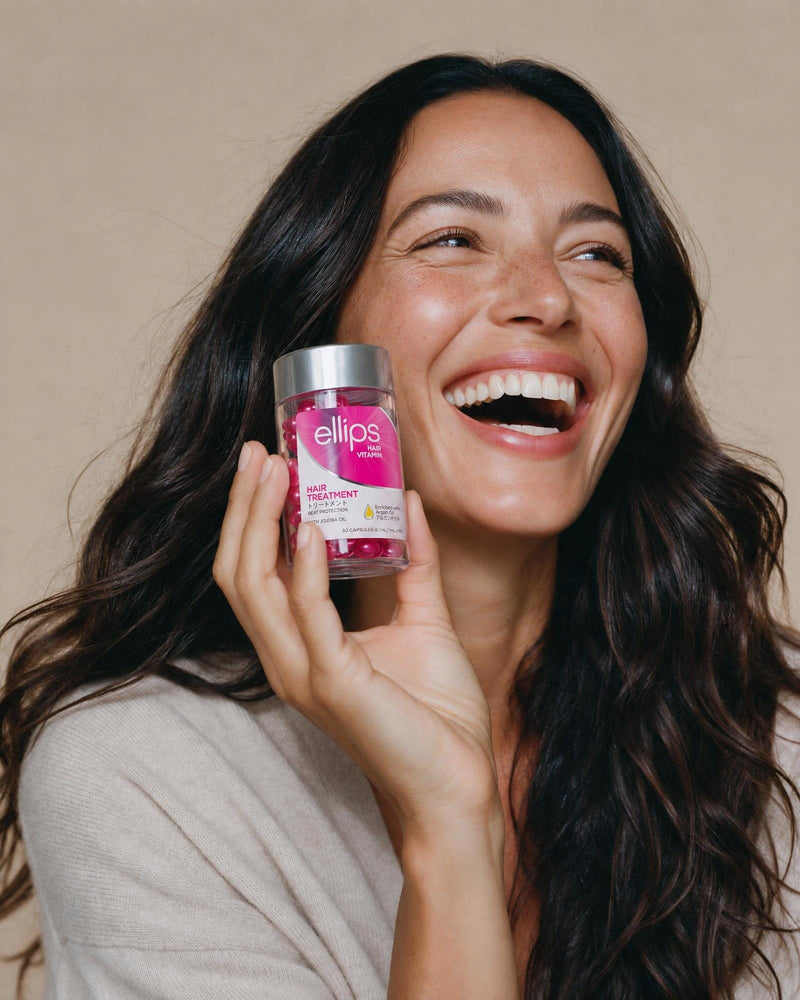 Naurava nainen, jolla on pitkät tummat hiukset, pitelee Ellips Hair Treatment -purkkia